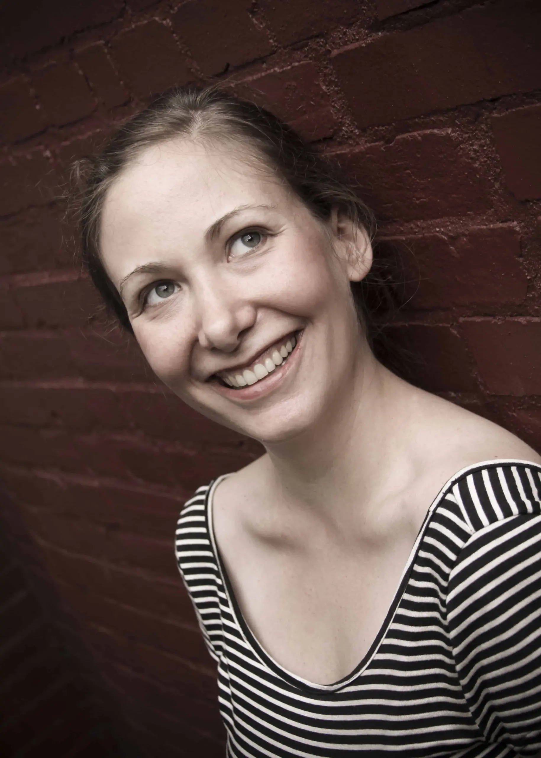 A woman with light skin and brown hair, wearing a black and white striped top, smiles while leaning against a red brick wall. She is looking slightly upward and to her left.
