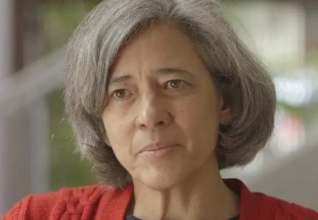 A middle-aged woman with gray hair and a thoughtful expression, wearing a red sweater, is looking slightly off-camera. The background is softly blurred.