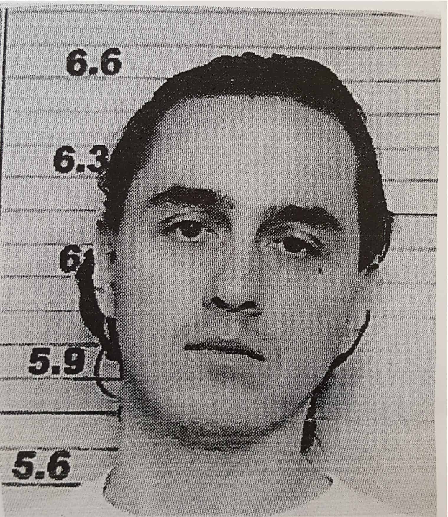 Black and white mugshot of a person with long dark hair, looking at the camera, standing in front of a height chart with measurements from 5.6 to 6.6 feet.