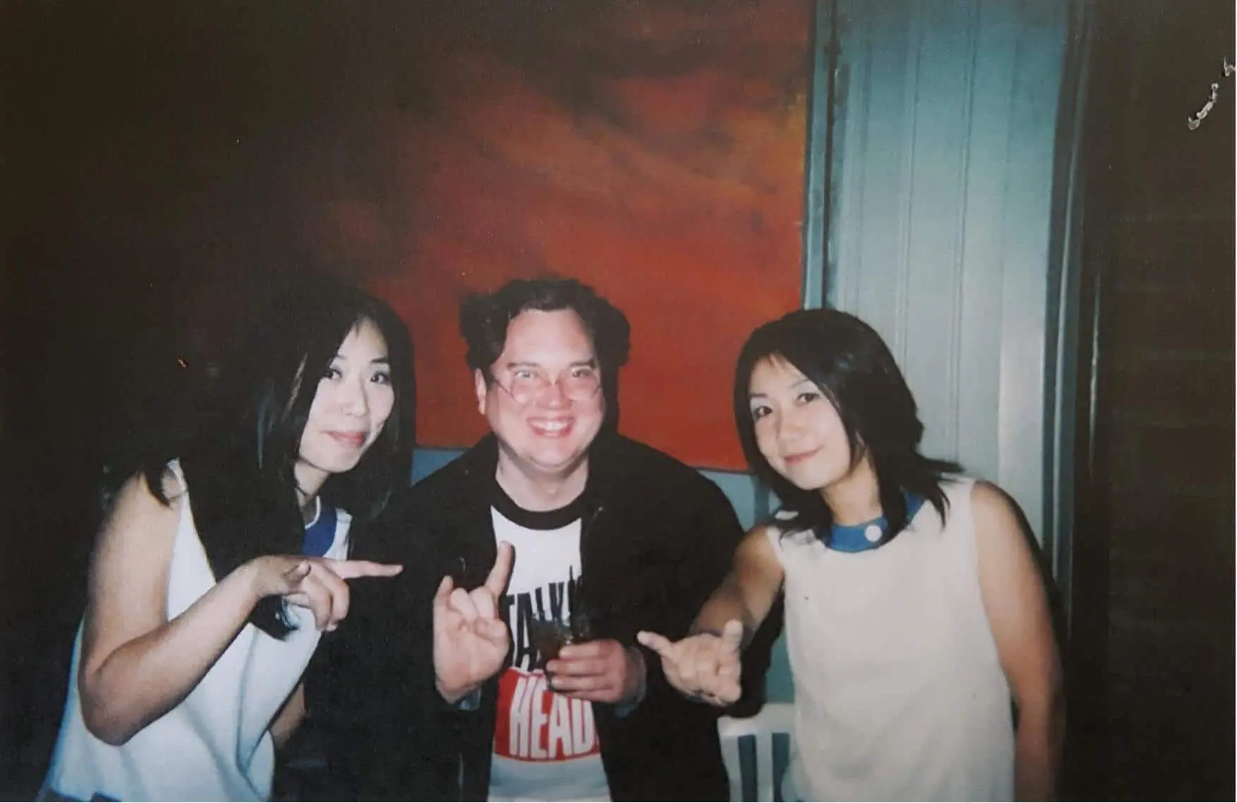 Three people smile at the camera; two women on either side make a rock on hand gesture, while the man in the center, wearing glasses and a band shirt, holds a drink. A red and orange painting hangs behind them.