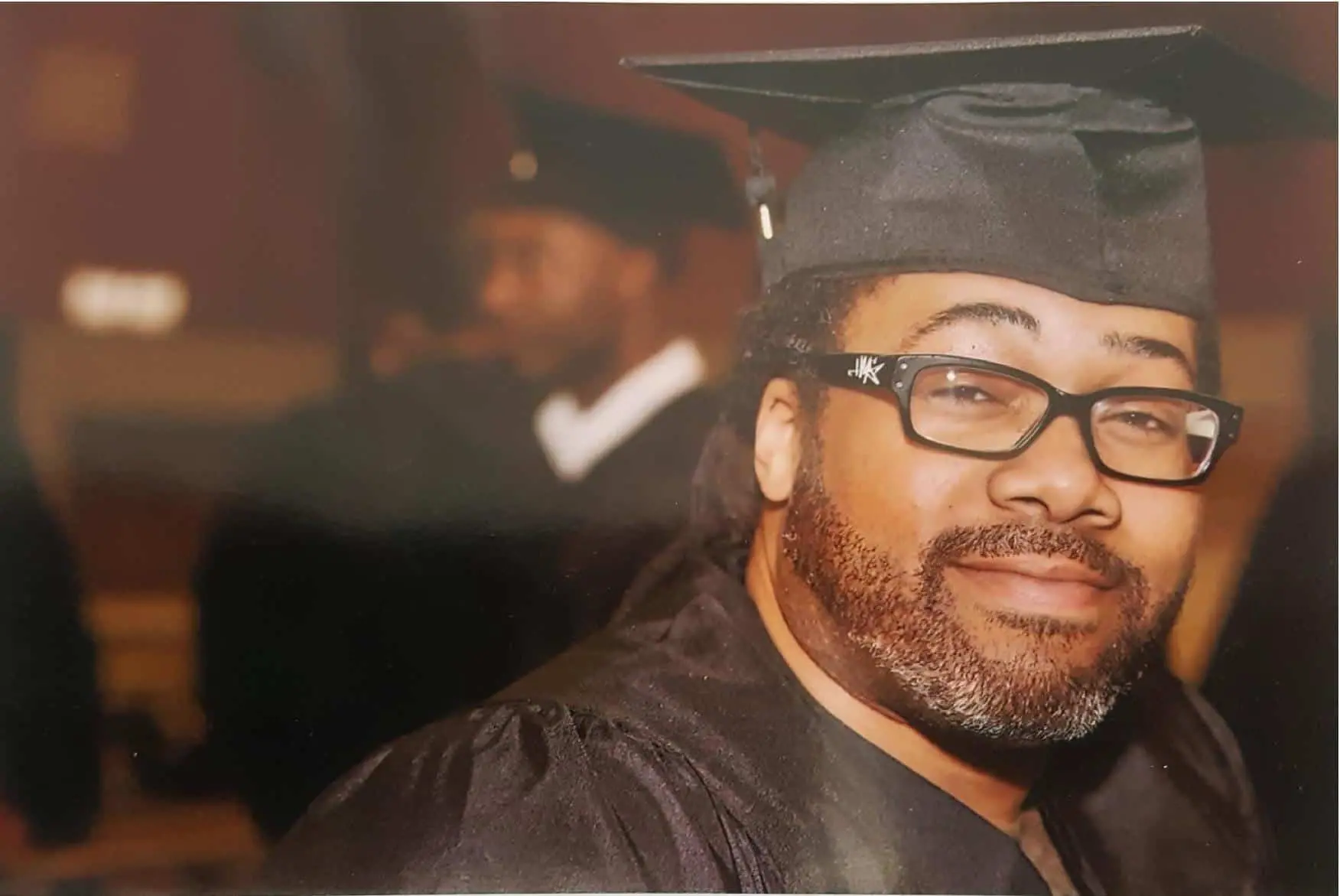A man wearing glasses and a graduation cap and gown smiles at the camera. The background is blurred with another graduate visible.