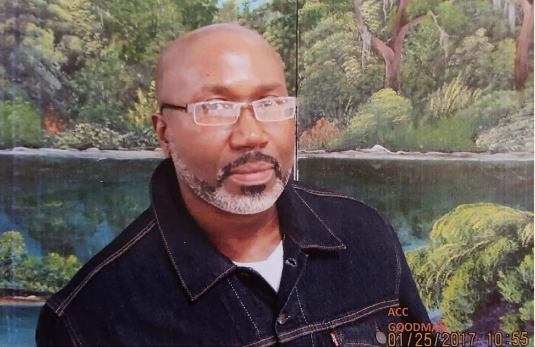 A man with glasses, a short beard, and a black denim jacket poses in front of a painted backdrop featuring trees and a river. The date on the photo is 01/25/2017.