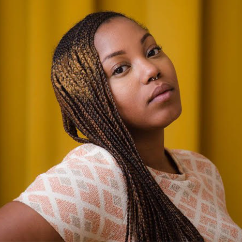 A woman with long, braided hair and a septum piercing looks confidently at the camera. She wears a light-patterned top and stands in front of a bright yellow background.