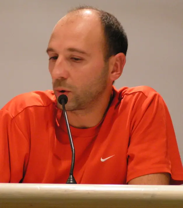 A man in an orange Nike t-shirt speaks into a microphone at a podium, looking down thoughtfully.