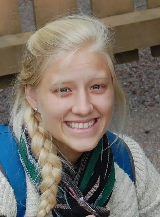 A young woman with blonde hair in a side braid smiles at the camera. She is wearing a striped scarf, a light sweater, and a blue backpack, with a wooden bench and gravel visible in the background.