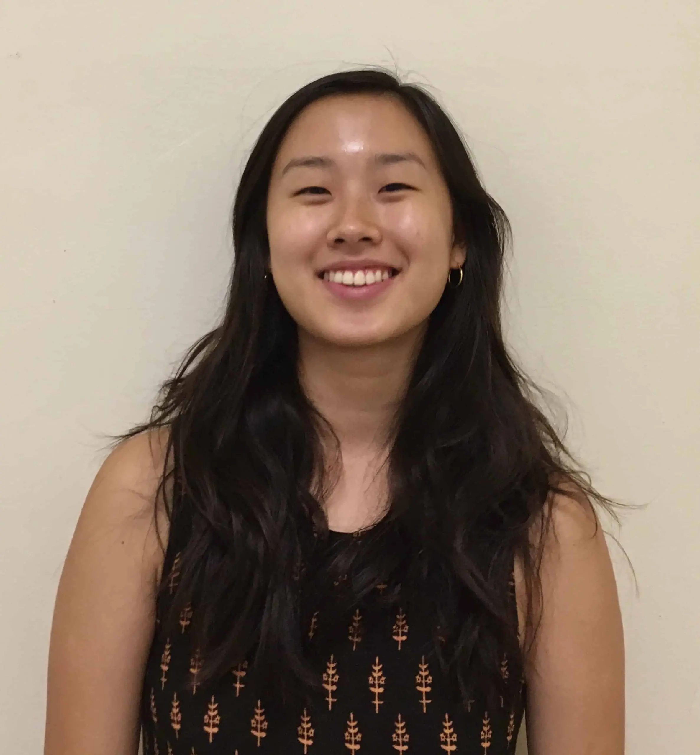 A young woman with long, dark hair and a sleeveless patterned top smiles while standing against a plain, light-colored wall.