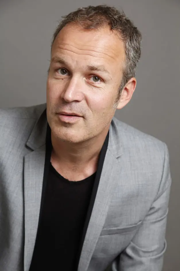 A middle-aged man with short, graying hair is wearing a light gray blazer over a black shirt, looking directly at the camera with a neutral expression against a plain gray background.
