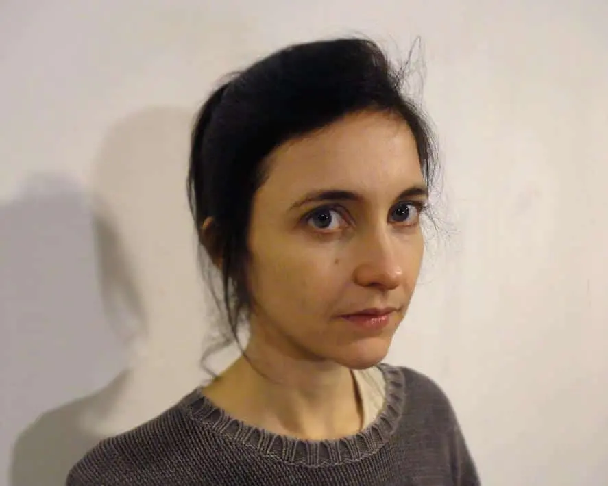 A woman with dark hair tied back, wearing a gray sweater, stands against a plain light-colored wall. She looks slightly to the side with a neutral expression.