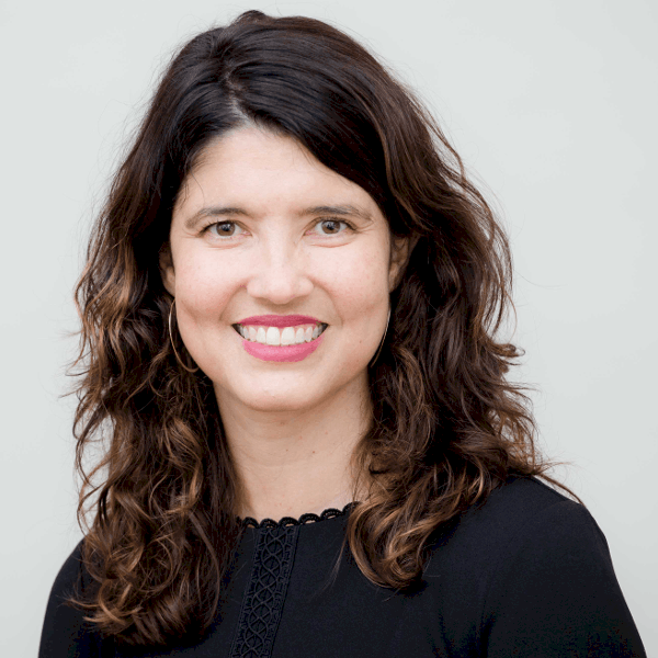 A woman with long, wavy brown hair smiles at the camera. She is wearing a black top and has a light, neutral background behind her.