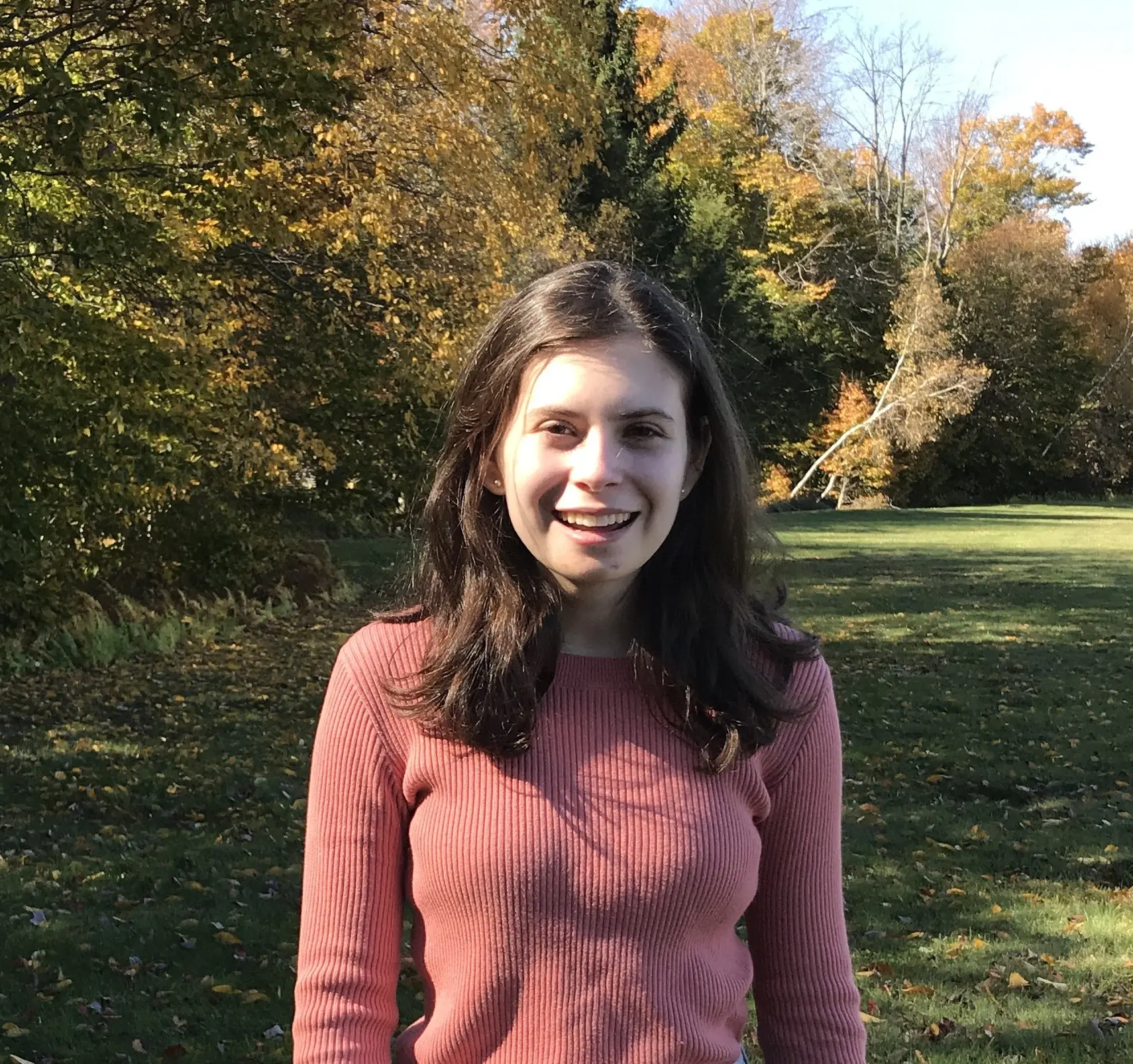 A young woman with long brown hair, wearing a pink ribbed sweater, stands smiling in a sunlit park with green grass and trees displaying autumn foliage in the background.