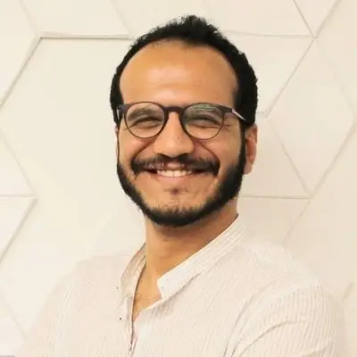 A smiling man with short curly hair, a beard, and glasses, wearing a light-colored shirt, stands in front of a white geometric-patterned wall.