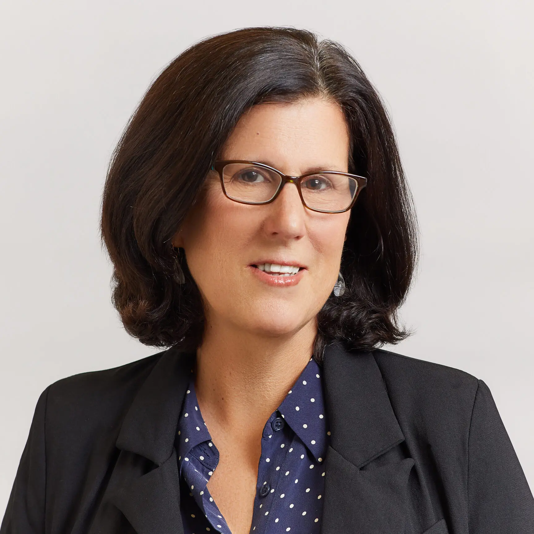 A woman with shoulder-length dark hair and glasses is wearing a black blazer over a navy blue blouse with white polka dots, smiling softly at the camera against a plain light background.