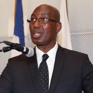 A man in a black suit, white shirt, and polka dot tie speaks into a microphone. He wears glasses and stands in front of a white and blue flag and a white background.