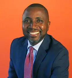 A smiling man in a navy blue suit, light blue shirt, and red patterned tie sits in front of an orange background.