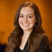A woman with long, wavy brown hair and a black blazer smiles at the camera against a warm, blurred orange background.