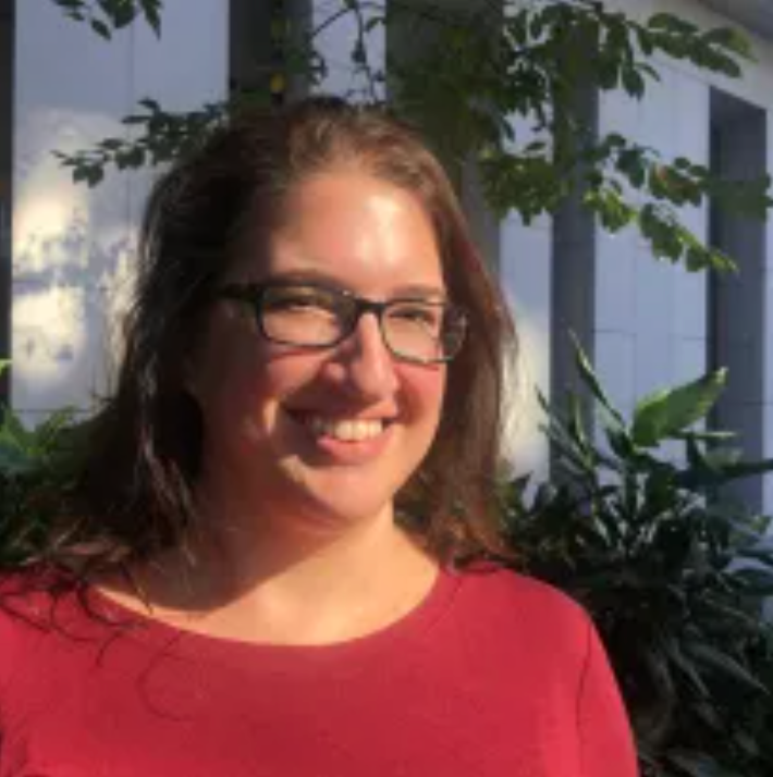 A woman with long brown hair and glasses, wearing a red top, smiles while standing outdoors in front of green plants and a building with white and dark vertical panels. Sunlight shines on her face.