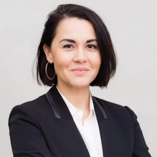 Woman with short dark hair, wearing a black blazer over a white shirt, smiles softly with arms crossed against a plain light background.