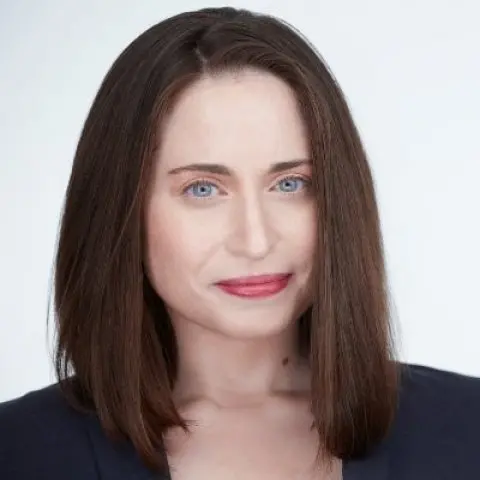 A woman with straight, shoulder-length brown hair and blue eyes, wearing a dark top, looks directly at the camera with a neutral expression against a light background.
