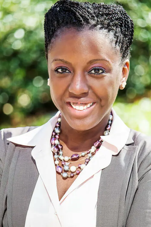 A woman with braided hair smiles confidently outdoors, wearing a gray blazer, light pink blouse, and a beaded necklace. The background is blurred greenery.