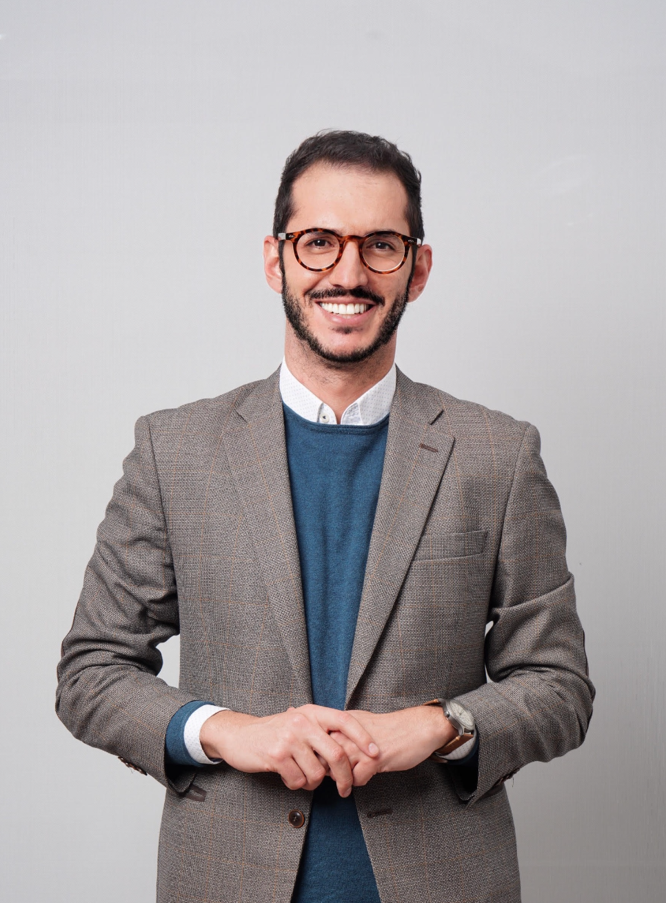 A man with short dark hair, a beard, and glasses, wearing a gray blazer over a blue sweater and white shirt, stands smiling with his hands loosely clasped in front of a plain light gray background.