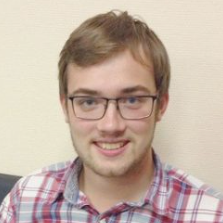 A young man with short brown hair and glasses smiles at the camera. He is wearing a red and white plaid shirt and sitting in front of a light-colored wall at a Global Press Freedom Advocacy Institute PEN America event.