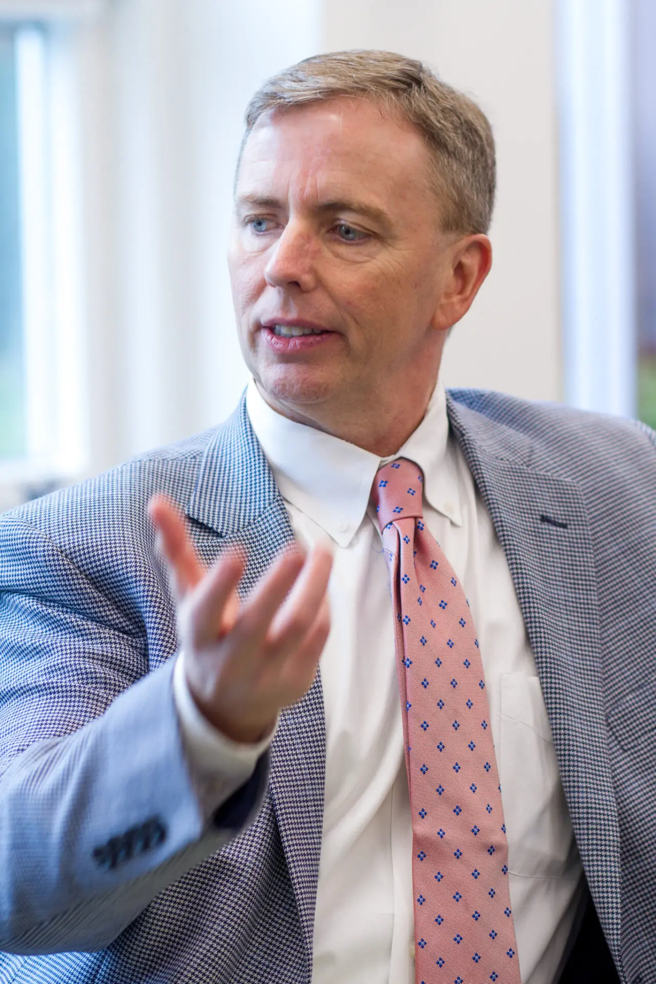 A man in a blue checked suit jacket, white shirt, and pink patterned tie gestures with his right hand while speaking, with a focused expression. The background is softly blurred with natural light coming through a window.