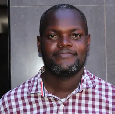 A man with short hair and a trimmed beard wearing a red and white checkered shirt stands in front of a dark tiled wall, looking at the camera with a slight smile.