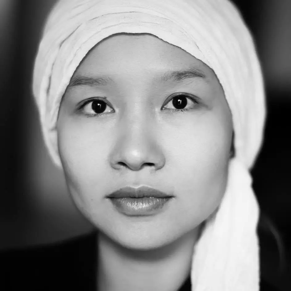 A person with a neutral expression wearing a light-colored headscarf gazes directly at the camera; the image is in black and white with a blurred background.