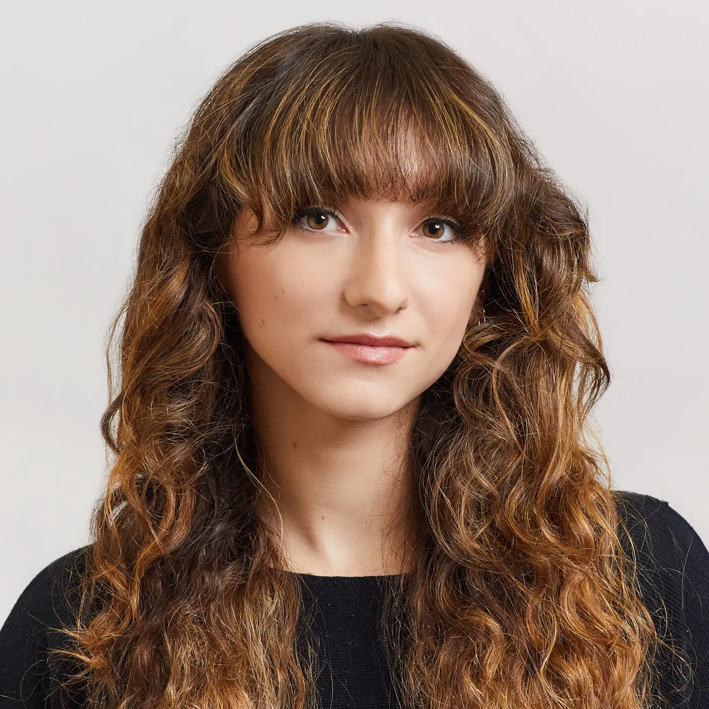 A young woman with long, wavy brown hair and bangs, wearing a black top, looks at the camera with a neutral expression against a plain light background.
