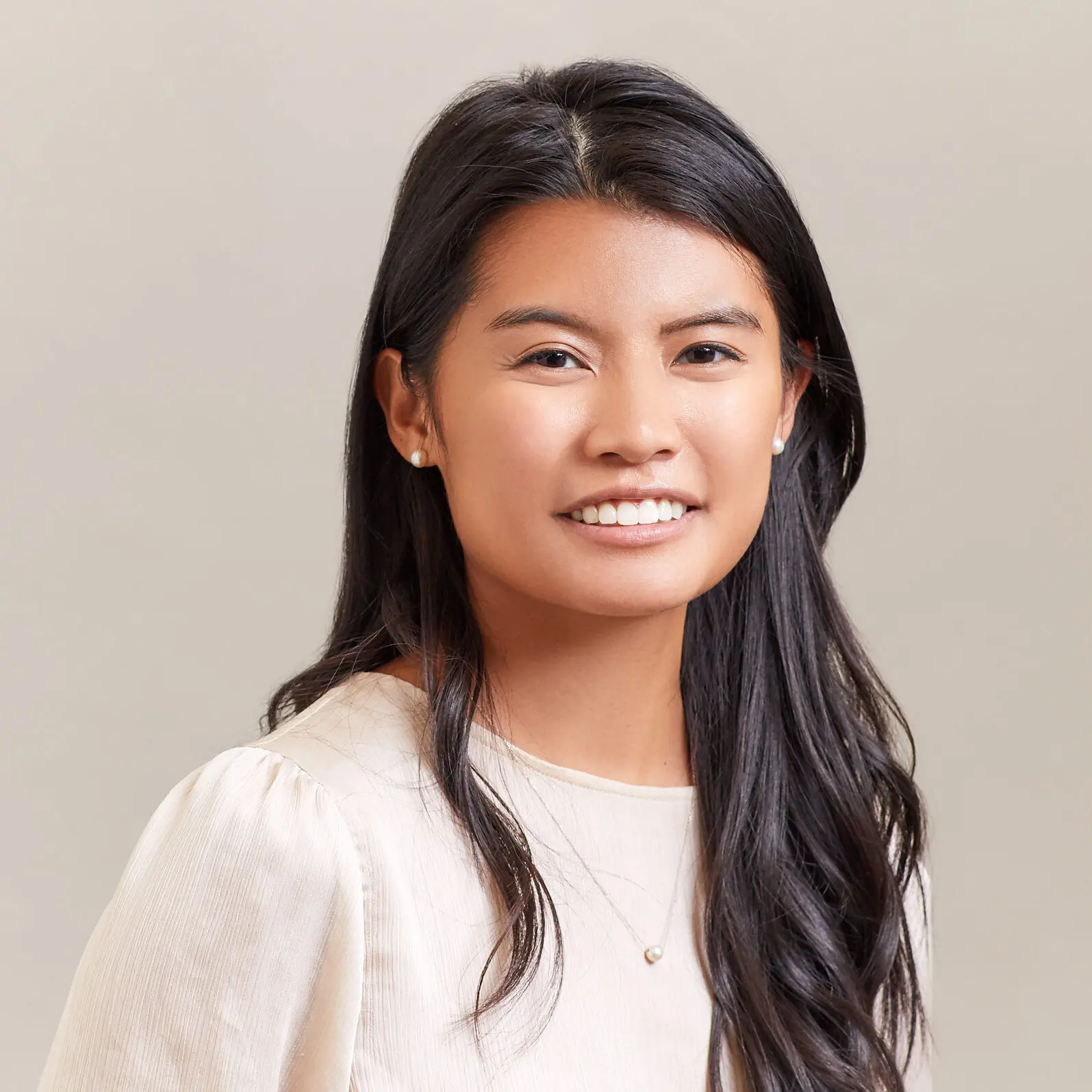 A woman with long dark hair wearing a light-colored blouse and a small necklace smiles at the camera against a plain, light background.