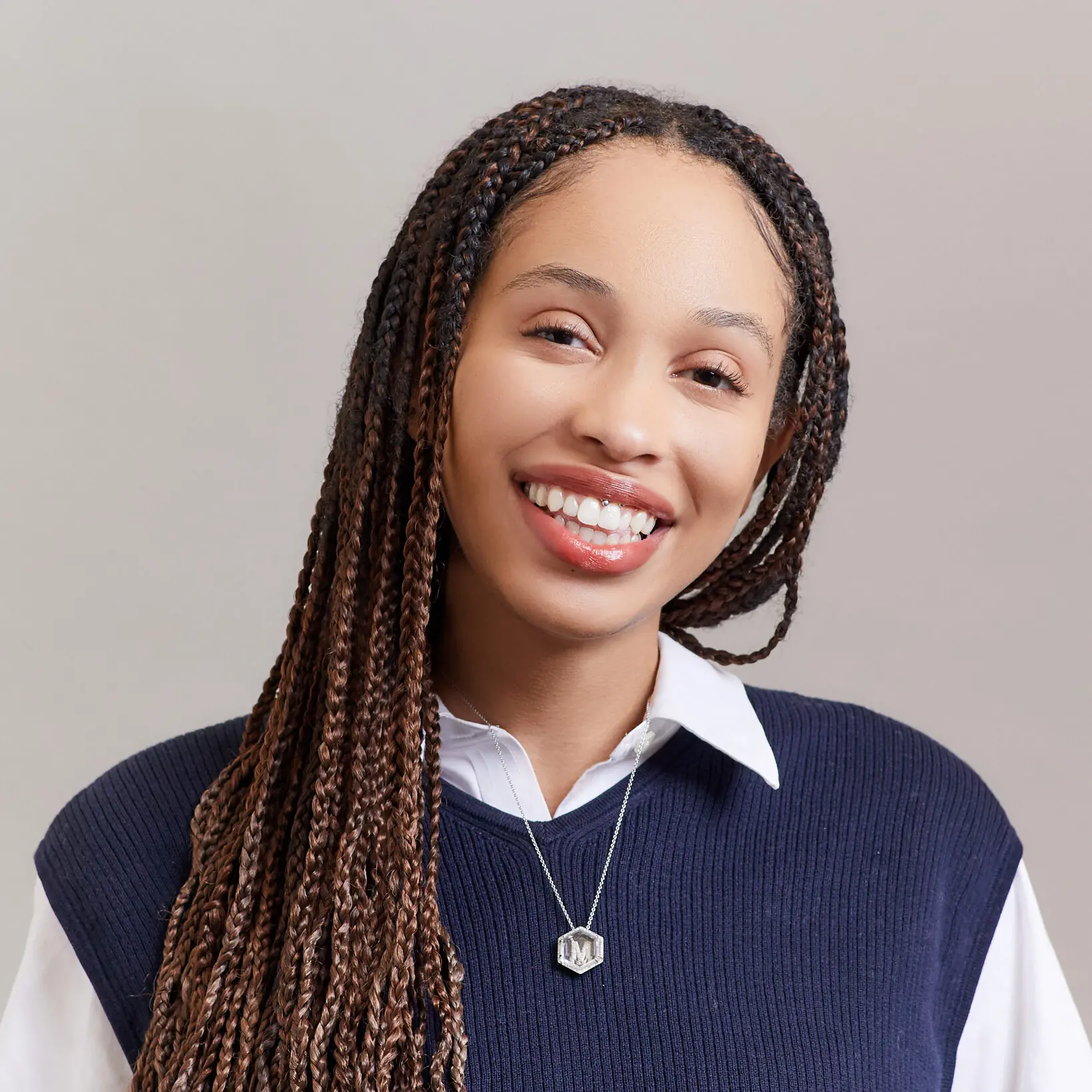 A smiling woman with long braided hair wears a navy sweater over a white collared shirt and a necklace with a pendant shaped like the letter M, standing against a plain light background.