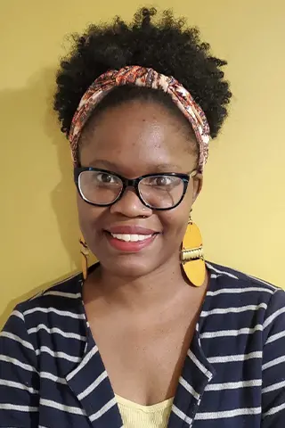 A woman with curly hair in a patterned headband, dark glasses, and large yellow earrings smiles at the camera. She wears a striped navy and white top over a yellow shirt, in front of a yellow background at a Pen America campus for all workshops.