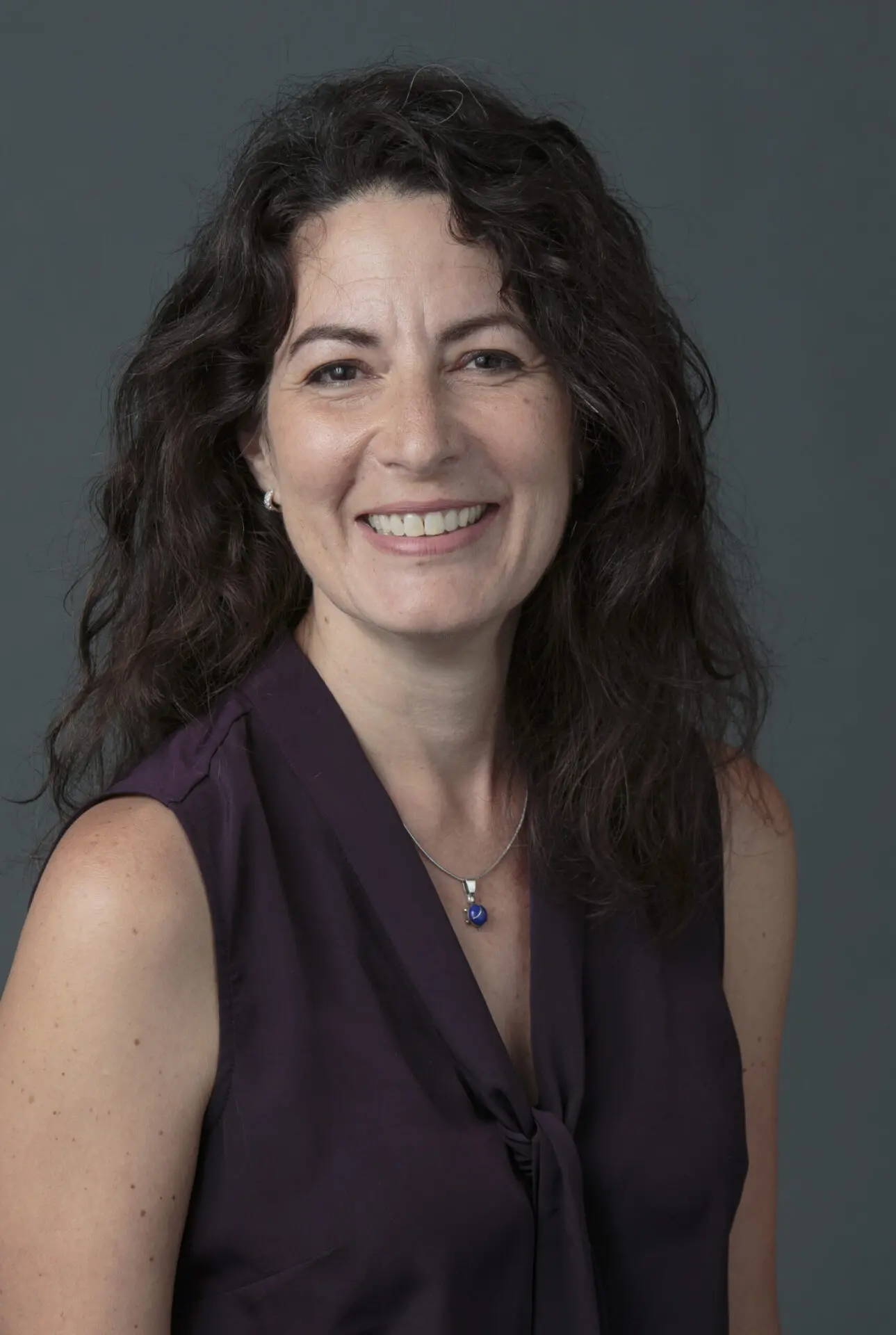 A woman with wavy dark hair smiles at the camera. Wearing a sleeveless purple blouse and a blue pendant, she poses against a plain dark background—ready to lead Pen America workshops and trainings on campus for all.