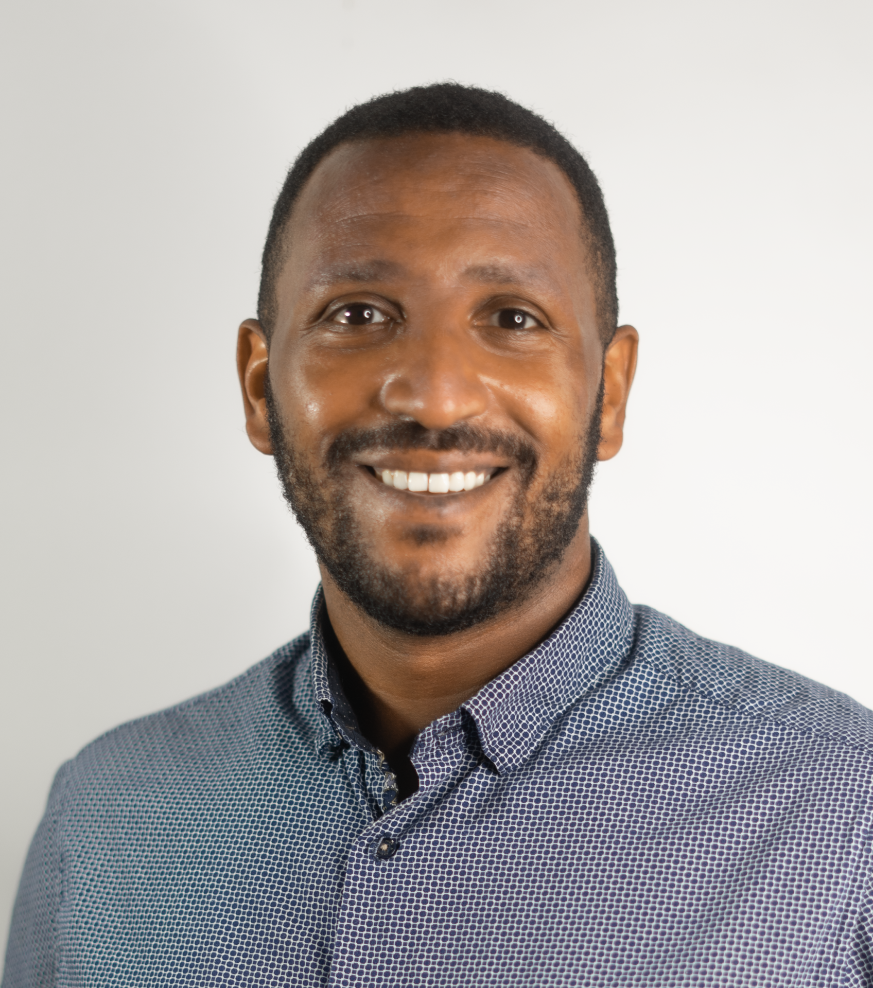 A man with short black hair and a beard, wearing a blue patterned shirt, smiles at the camera against a plain, light-colored background.