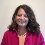 A woman with curly brown hair smiles at the camera. She is wearing a bright pink jacket, a white top, a silver necklace, and small hoop earrings. The background is a plain, light-colored wall.