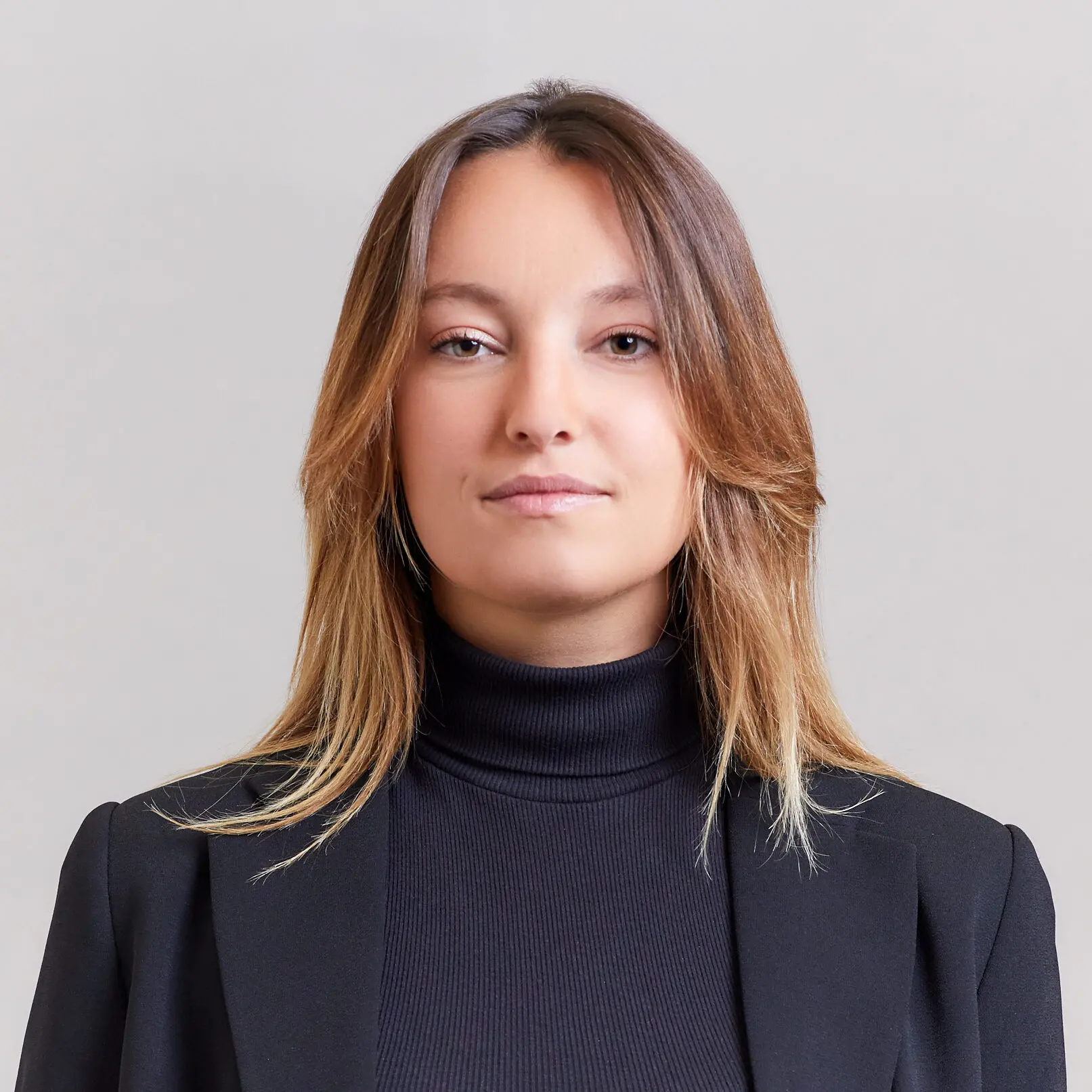 A woman with straight, light brown hair wearing a black turtleneck and black blazer stands against a plain, light gray background, looking confidently at the camera.
