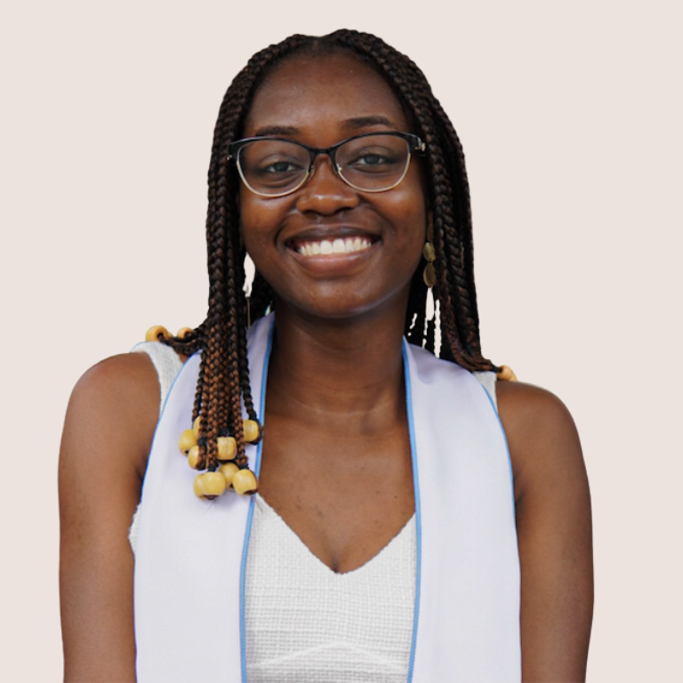 A young woman with glasses and long braided hair, wearing a white dress and a light blue graduation stole, smiles at the camera against a plain light background.