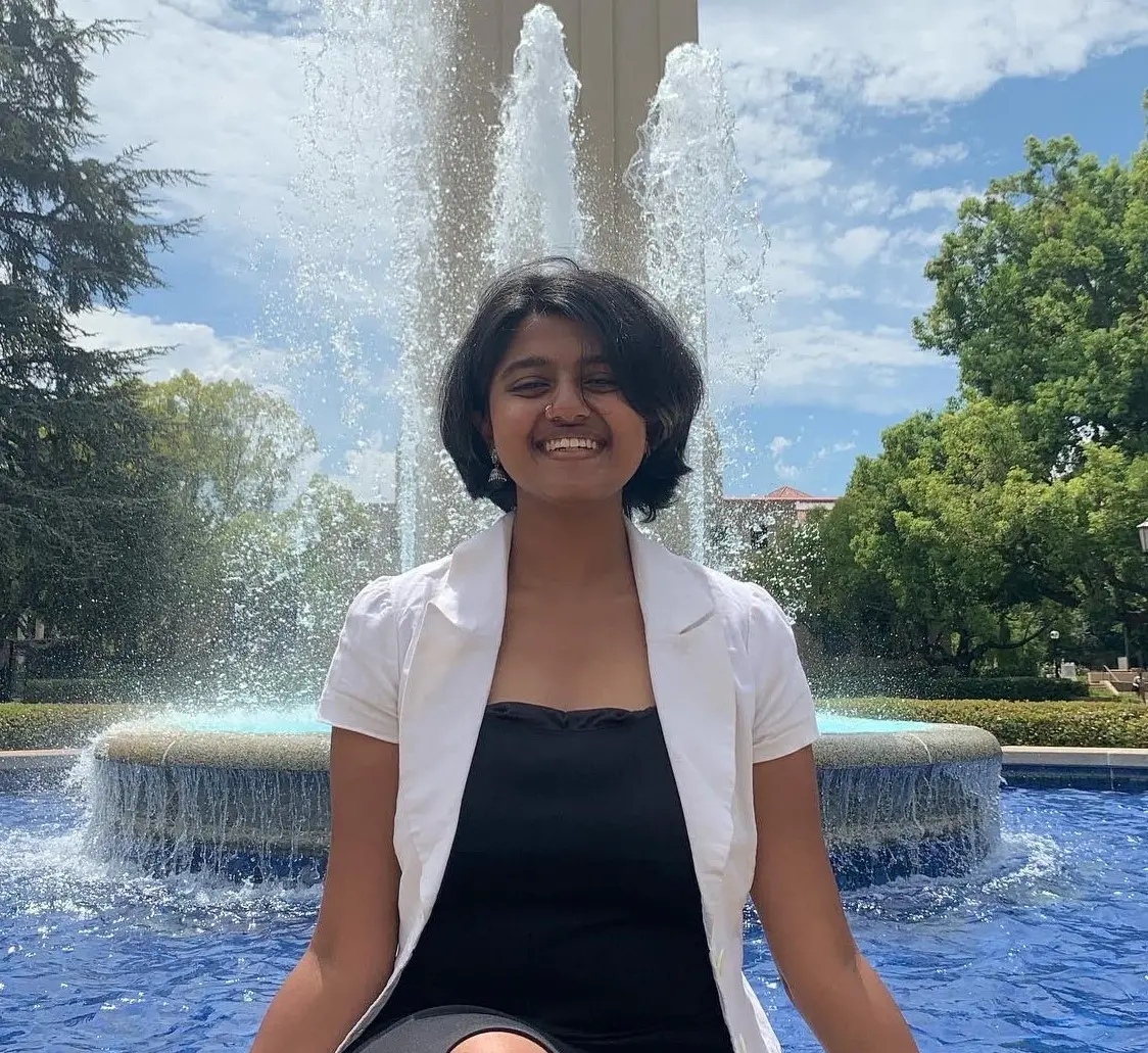 A person with short dark hair sits smiling in front of a fountain, wearing a white blazer over a black top. Green trees and a blue sky with clouds are visible in the background.
