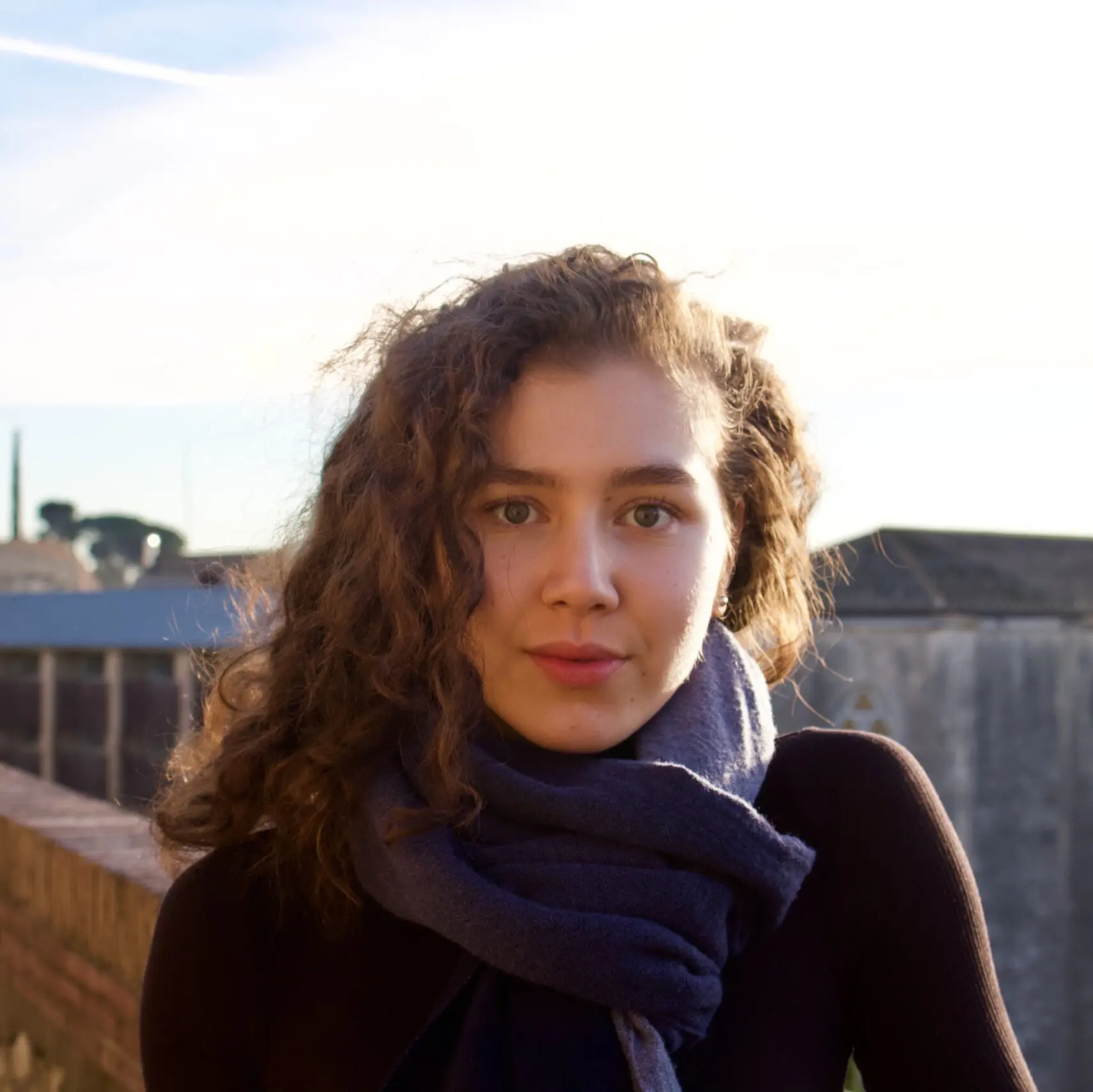 A young woman with curly brown hair and a dark scarf stands outdoors, looking at the camera. The background shows a bright sky, some buildings, and a brick wall.