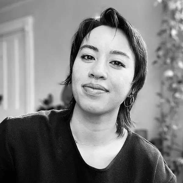 Black and white portrait of a person with short dark hair, hoop earrings, and a nose ring, wearing a dark top, smiling slightly while looking at the camera. Blurred indoor background with a plant visible, capturing themes of online abuse and digital safety.