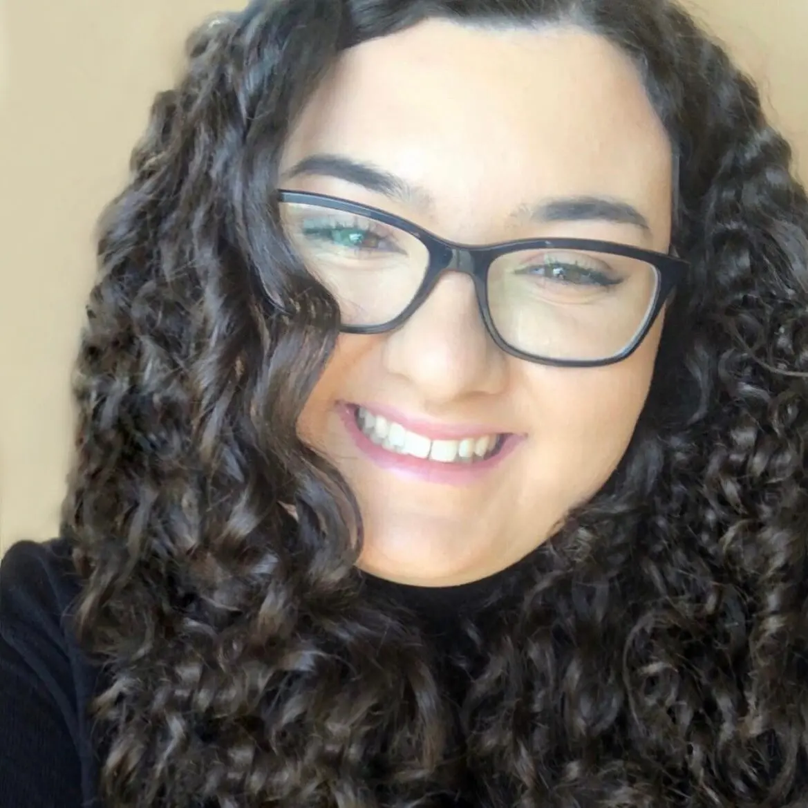 A person with long, dark, curly hair and glasses smiles at the camera. They are wearing a black top against a soft, neutral background, radiating hope and resilience in the face of prison and justice challenges.