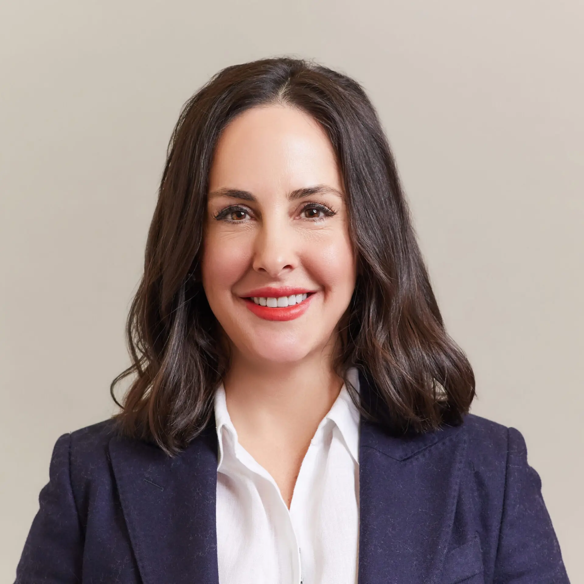 A woman with shoulder-length dark hair smiles at the camera. She is wearing a white blouse and a dark blazer, standing against a plain light-colored background.