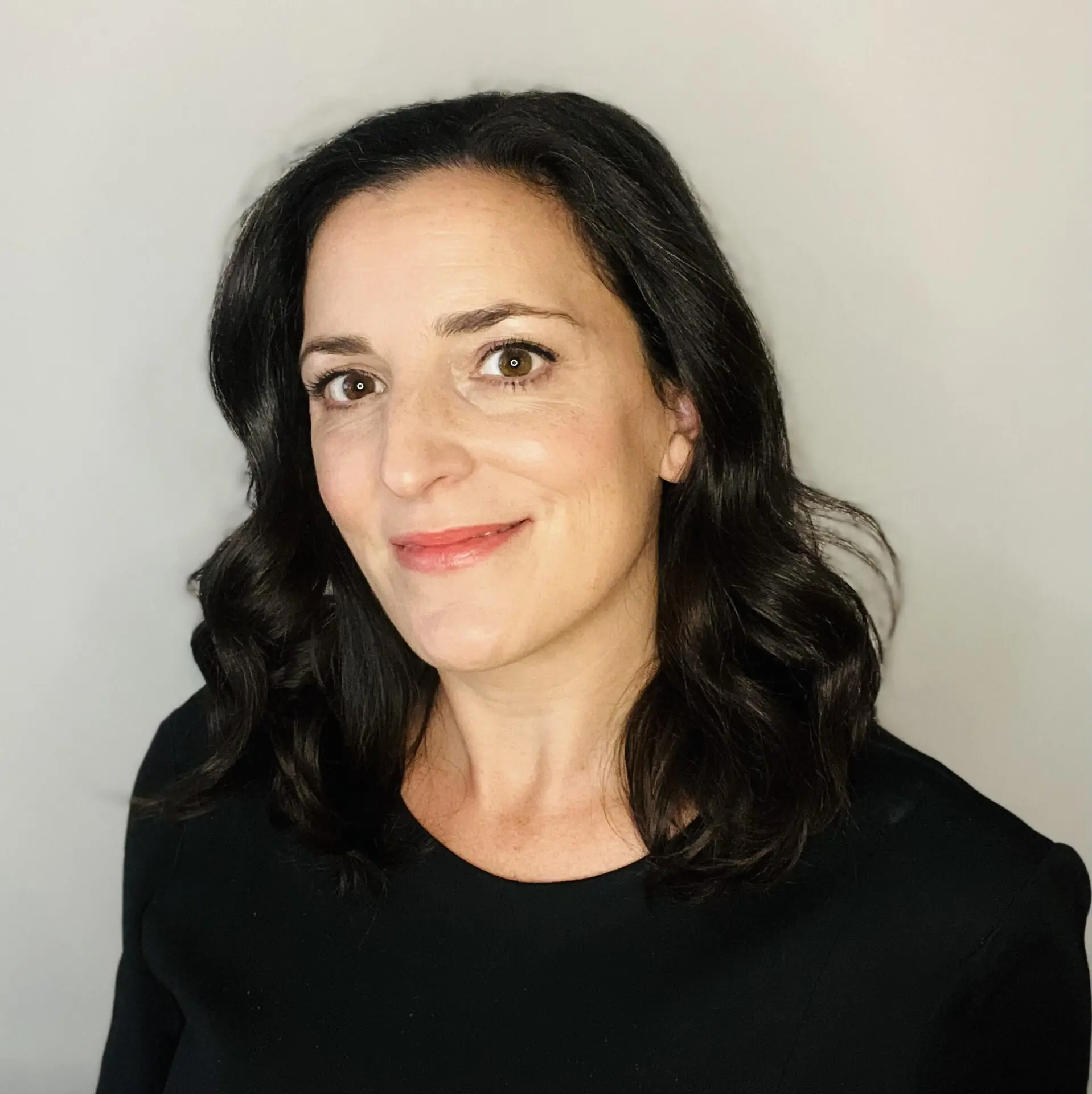A woman with wavy dark hair and fair skin smiles slightly, wearing a black top, against a plain light gray background.