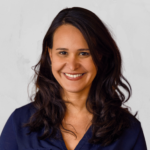 A woman with long, wavy dark hair and a big smile is wearing a navy blue shirt, standing in front of a light, plain background—her confidence mirroring the spirit celebrated in the Freedom to Write Index 2022.