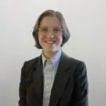 A person with short brown hair, wearing glasses, a light blue button-up shirt, and a dark blazer, smiles at the camera against a plain light-colored background.