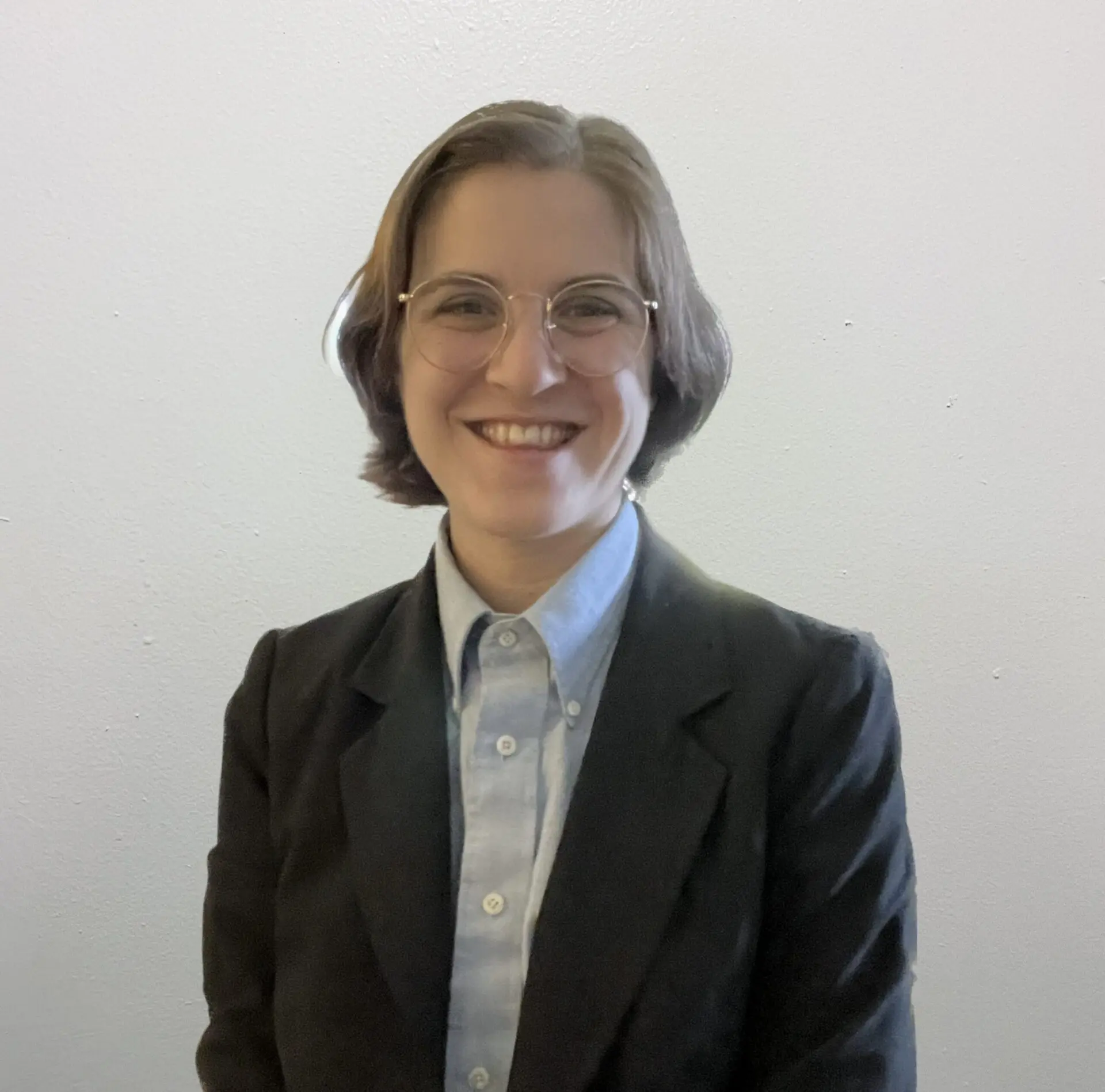 A person with short brown hair, wearing glasses, a light blue button-up shirt, and a dark blazer, smiles at the camera against a plain light-colored background.