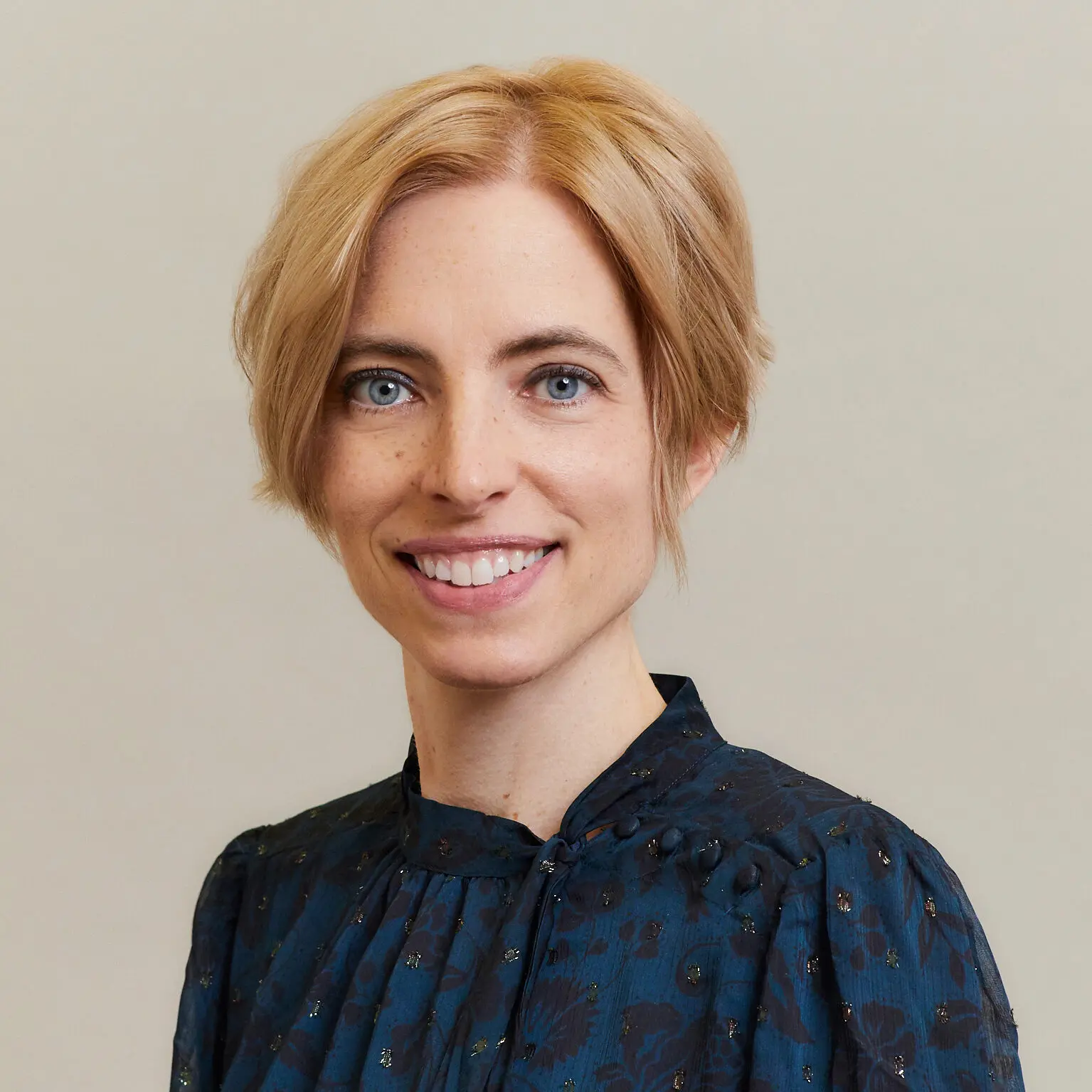 A woman with short blonde hair and blue eyes smiles at the camera. She is wearing a dark blue patterned blouse and is posed against a plain light background.
