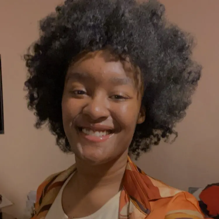 A person with a large, curly afro smiles at the camera. They are wearing a patterned shirt with shades of orange and brown over a white top. The background is softly lit and neutral.