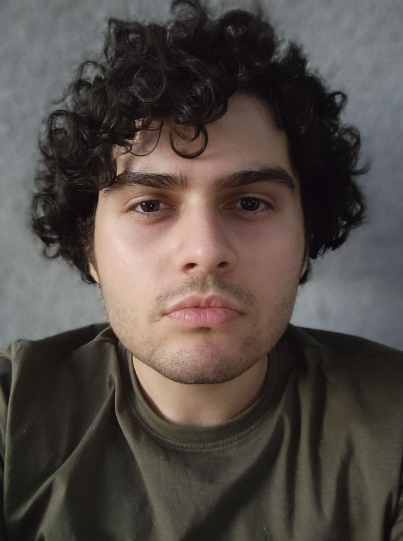 A person with short, curly dark hair and a trimmed beard looks directly at the camera with a neutral expression, wearing a green shirt, against a plain, gray background.