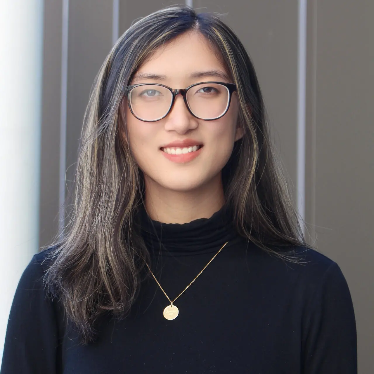 A person with long, straight brown hair and glasses is smiling, wearing a black turtleneck top and a gold necklace, standing in front of a neutral-colored background.